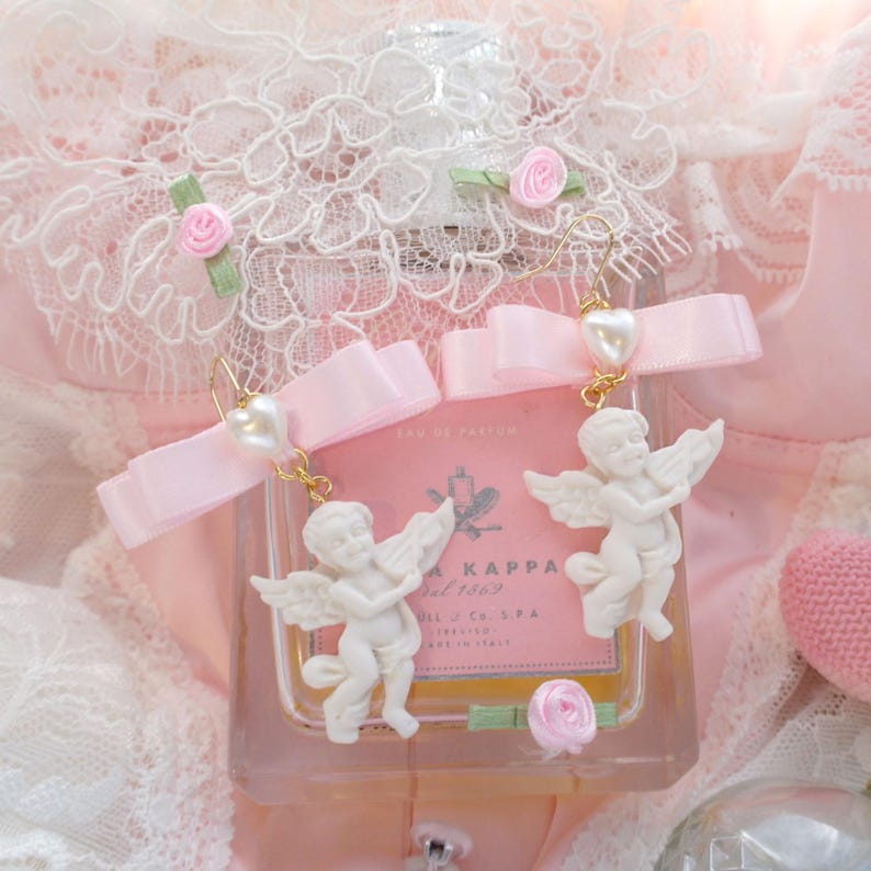 K&ouml;nnte beinhalten: Wei&szlig;e Porzellan-Engelohrringe mit rosa Schleifen und Perlenakzenten. Die Ohrringe h&auml;ngen an einer goldenen Kette.