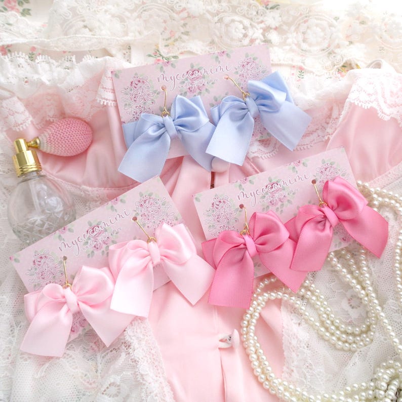 May include: Two pairs of earrings with large satin ribbon bows. One pair is light blue and the other is pink. The earrings are displayed on a white background with floral designs and the text "myceenhime".