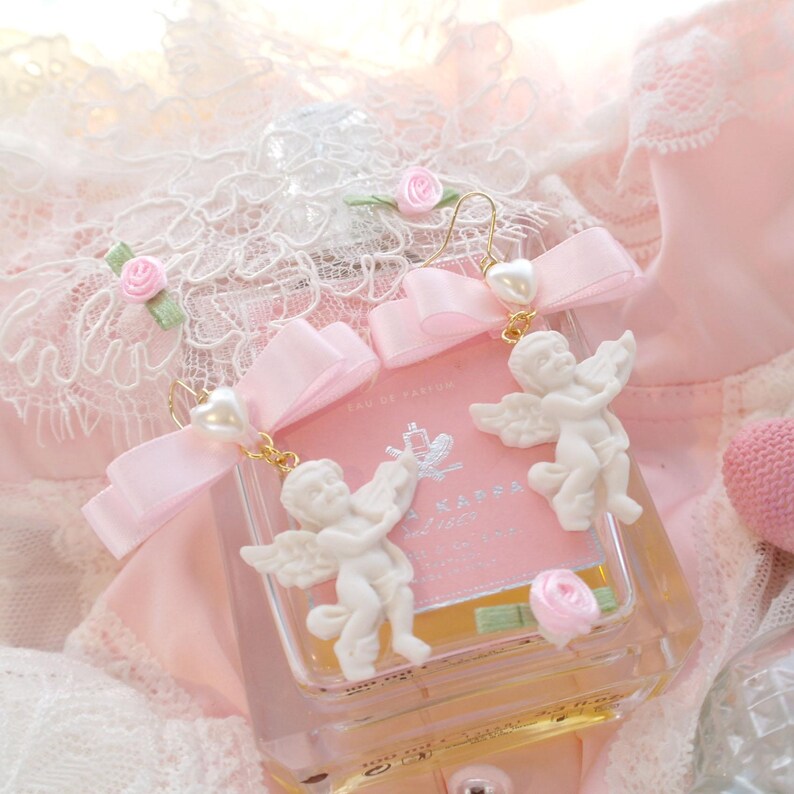 K&ouml;nnte beinhalten: Ein Paar zarte Ohrringe mit einem wei&szlig;en Cherub-Anh&auml;nger, einer rosa Schleife und einem Perlenakzent. Die Ohrringe werden auf einer rosa Flasche mit einem wei&szlig;en Etikett pr&auml;sentiert.