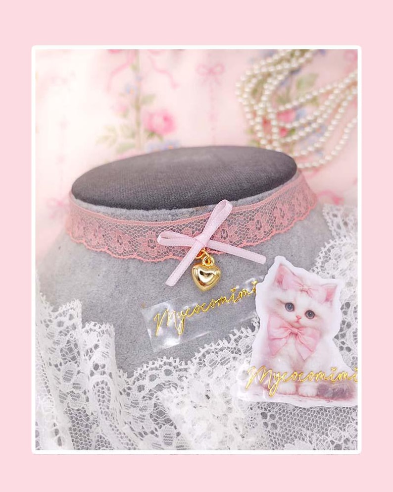 May include: A pink lace choker necklace with a gold heart pendant and a small bow. A sticker of a white kitten with a pink bow is next to the necklace. The background includes a pearl necklace and a floral pattern.