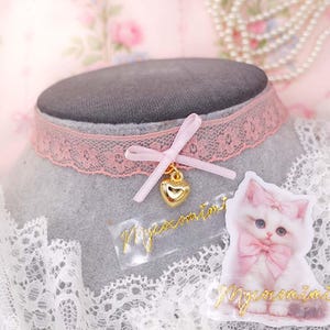 May include: A pink lace choker necklace with a gold heart charm and a pink bow. A sticker of a white kitten with a pink bow is next to the choker. The text "Mycocomini" is on the sticker and the choker.