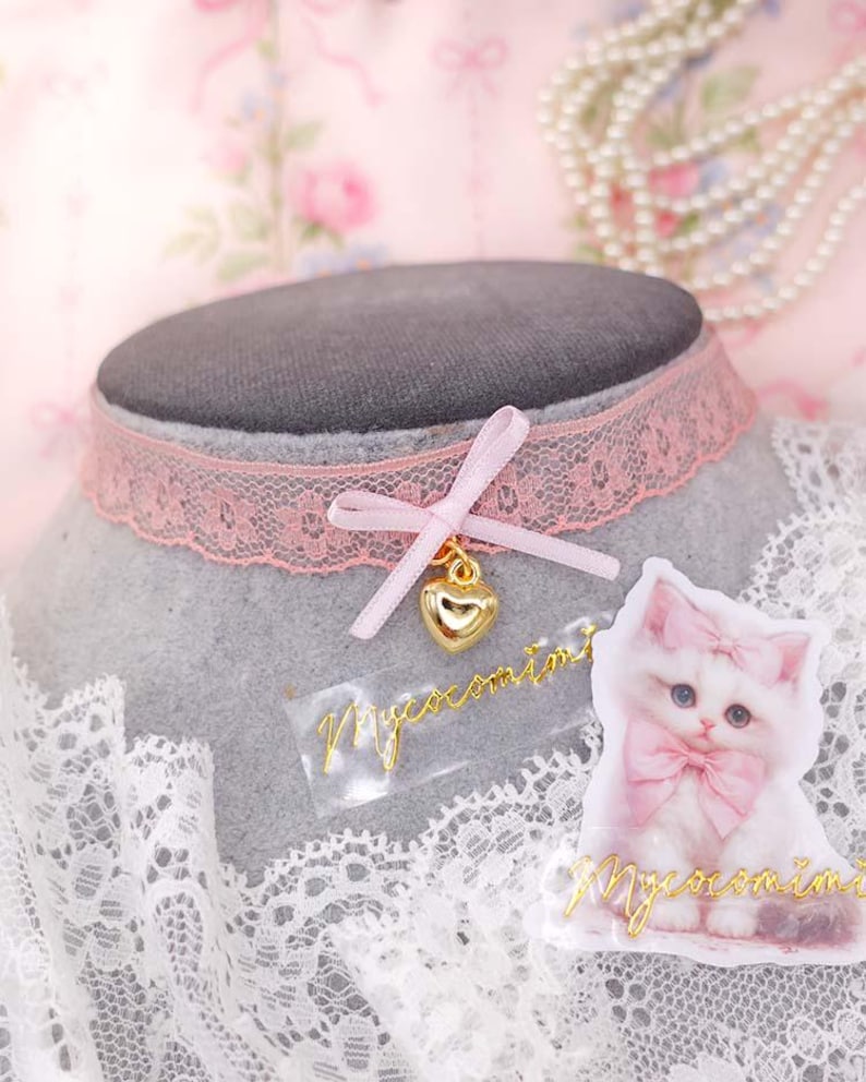 May include: A pink lace choker necklace with a gold heart charm and a pink bow. A sticker of a white kitten with a pink bow is next to the choker. The text "Mycocomini" is on the sticker and the choker.