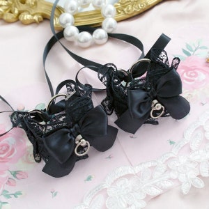 May include: A pair of black lace cuffs with satin bows and silver-toned metal rings. The cuffs are adorned with delicate lace and black satin bows. Black ribbons extend from the cuffs. The background includes a pearl necklace and a decorative mirror.