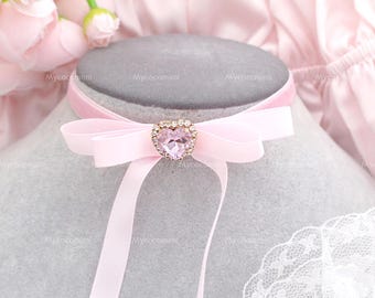 Peut inclure: Un collier ras du cou rose clair avec un pendentif en forme de cœur. Le pendentif est orné de petits cristaux transparents autour d'une grosse pierre rose en forme de cœur. Le collier a un nœud et de longs rubans.