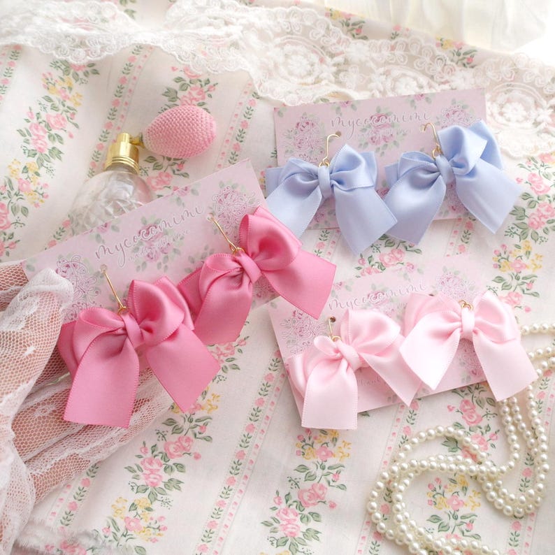 May include: Three pairs of pink and light blue satin ribbon bow earrings. The earrings are on a white background with floral patterns. The earrings are labeled "mycooshine".