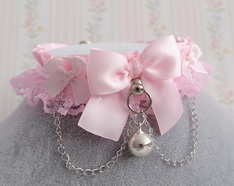 May include: A pink choker necklace with a large satin bow and a smaller bow on each side. The choker has a lace trim and a silver bell charm hanging from a chain. The necklace is designed with a feminine aesthetic.