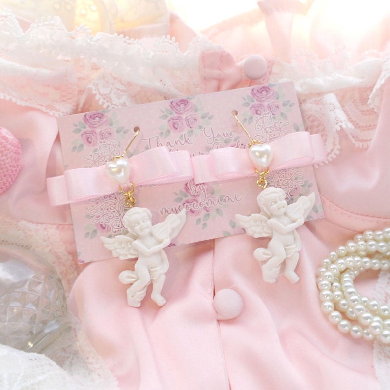K&ouml;nnte beinhalten: Ein Paar wei&szlig;e Cherub-Ohrringe mit Perlenakzenten. Die Ohrringe werden auf einer rosa Karte mit dem Text "Thank You" und "by mysecretmini" pr&auml;sentiert.