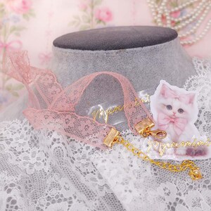 May include: A pink lace choker necklace with gold-colored clasps and a delicate gold chain. A small, illustrated cat with a pink bow is next to the necklace. The word "mignon" is written in gold script.