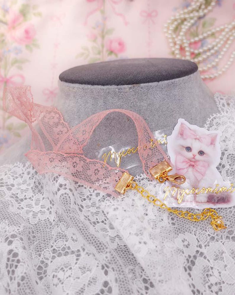 May include: A pink lace choker necklace with gold-colored clasps and a delicate gold chain. A small, illustrated cat with a pink bow is next to the necklace. The word "mignon" is written in gold script.
