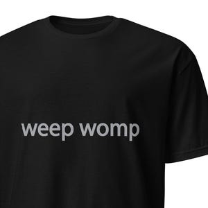 Puede incluir: Camiseta negra con la frase "weep womp" impresa en gris claro en el pecho. La camiseta tiene mangas cortas y cuello redondo. La tela parece ser un material suave y cómodo.