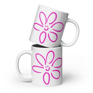 May include: Two white ceramic mugs stacked on top of each other. Each mug features a large, pink, cartoon-style flower design with a smiling face in the center. The mugs have a curved handle and a glossy finish.