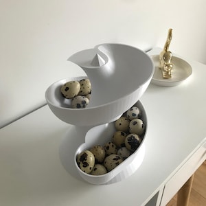 May include: A white, spiral-shaped egg holder filled with speckled quail eggs. The modern design features two curved bowls connected by a central support. A gold cat figurine sits on a small, round dish in the background.