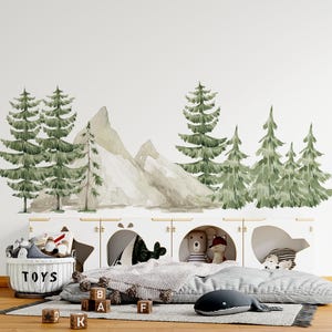 May include: Watercolor wall decal featuring a forest scene with green pine trees and gray mountains. A white storage unit with open compartments holds toys, a whale-shaped pillow, and a gray blanket. Wooden blocks spell out letters.