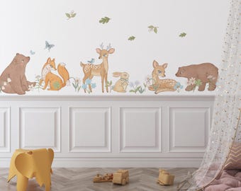 May include: A set of wall decals featuring illustrations of a brown bear, an orange fox, a spotted deer, a small rabbit, and a second brown bear. The animals are surrounded by blue flowers, green leaves, and a small blue bird, all arranged in a horizontal line.