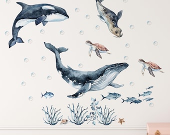 May include: A set of watercolor wall decals featuring a black and white orca, a gray seal, a large blue whale, two sea turtles, several small fish, blue coral, a seashell, a starfish, and scattered bubbles arranged across a plain white surface.