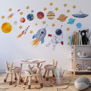 May include: A set of watercolor-style wall decals featuring space-themed elements including an astronaut, rockets, a flying saucer, various planets, and yellow stars. The designs are arranged on a wall above a small round table and chairs.
