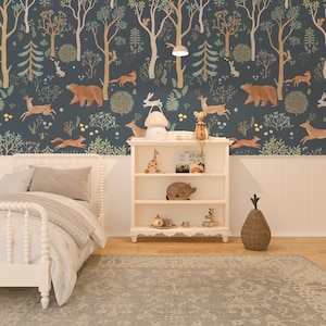 May include: A white wooden bed frame with a white headboard and footboard. The bed is made with a white duvet cover and a beige throw blanket. The bed is in a room with a dark blue wallpaper featuring a woodland scene with animals, trees, and flowers. A white bookshelf with three shelves is to the right of the bed. The bookshelf is decorated with a giraffe figurine, a small pig figurine, a teacup and saucer, and a stack of books. A wicker pineapple-shaped basket is on the floor next to the bookshelf.