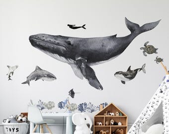 May include: A set of watercolor-style wall decals featuring a large humpback whale, a shark, an orca, a sea turtle, a manta ray, and a diver. The marine life illustrations are rendered in shades of gray and black, accompanied by blue coral accents.