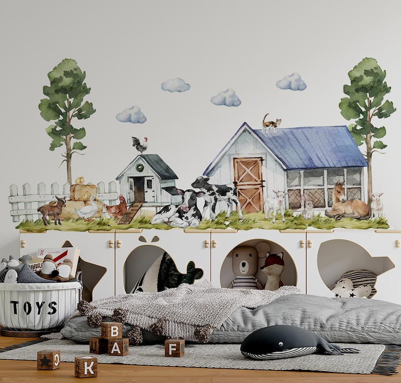 May include: A white wall with a farm scene decal featuring a barn, a chicken coop, cows, goats, a cat, and a horse. The decal is above a white shelf with three cubbyholes. The cubbyholes are decorated with a star, a fish, and a bear. There are toys and blocks on the floor in front of the shelf.