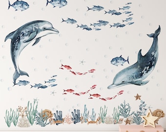 May include: A set of wall decals featuring two large blue dolphins, schools of smaller blue and red fish, scattered bubbles, and a bottom border of green seaweed, coral, and crabs. The aquatic elements are rendered in a watercolor style.