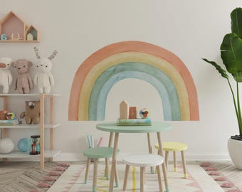 May include: A large watercolor-style rainbow wall decal featuring muted orange, yellow, and blue arches. The decal is positioned above a round light green table with three small stools, a white shelf with plush toys, and a tall green potted plant.