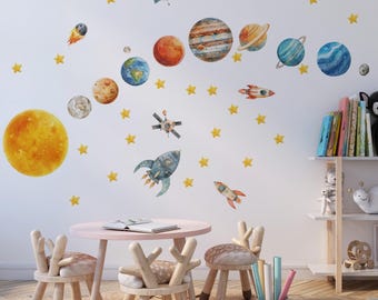 May include: A set of watercolor-style wall decals featuring a large yellow sun, various planets including Earth, Saturn, and Jupiter, three different rockets, two satellites, and numerous small yellow stars arranged across a white wall.