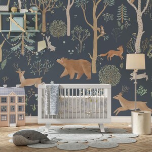 May include: A white cot with a blue and white patterned bedding set in a nursery with a blue and green woodland mural featuring a bear, deer, fox, and rabbit. A white lamp with a cream shade and a brown toy house are also in the room.
