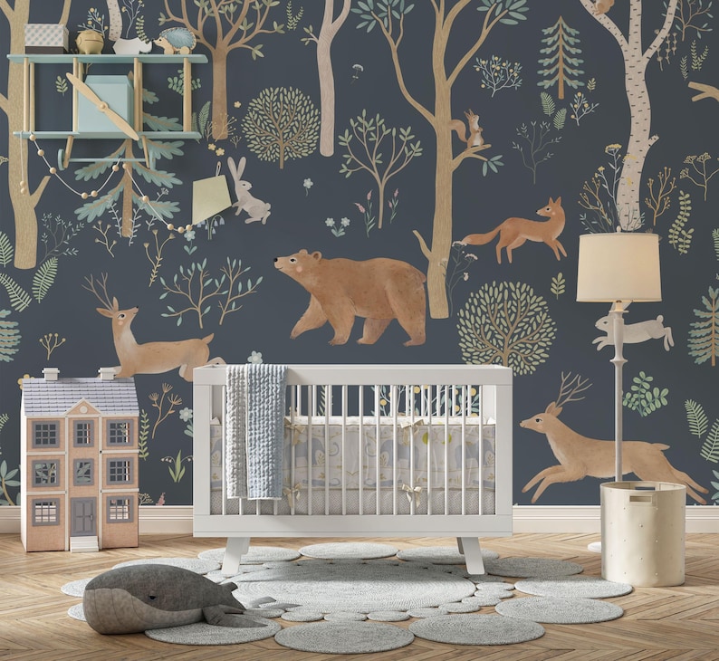 May include: A white cot with a blue and white patterned bedding set in a nursery with a blue and green woodland mural featuring a bear, deer, fox, and rabbit. A white lamp with a cream shade and a brown toy house are also in the room.
