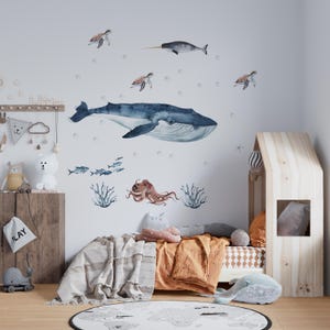 May include: A set of wall decals featuring a large blue whale, a narwhal, sea turtles, an octopus, small fish, and coral. The watercolour-style illustrations are arranged on a wall above a bed, depicting various marine life in shades of blue, brown, and grey, creating an underwater scene for a room.