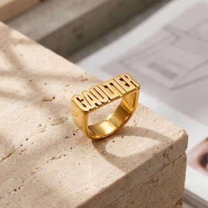 Peut inclure: Une bague dorée avec le nom "GAUTIER" en lettres majuscules. La bague est posée sur une surface texturée et claire, avec un arrière-plan flou comprenant un magazine. Un bijou audacieux et affirmé.
