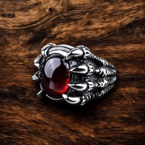 Puede incluir: Un anillo de plata con una piedra preciosa roja engastada en un diseño de garra. El anillo está hecho para parecer una garra de dragón.