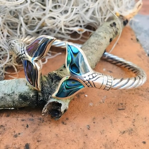 Puede incluir: Una pulsera de plata con dos abalorios de cola de sirena de concha de abulón. La pulsera tiene un diseño de cuerda trenzada.