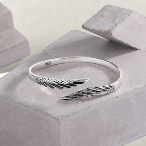 May include: Silver bangle bracelet with a wing design. The bracelet is open and has a textured finish.