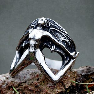 Op de afbeelding: Een zilverkleurige ring met een gestileerde figuur met uitgestrekte armen en lang haar. Het ontwerp bevat vleermuisachtige vleugels en een hartvormige uitsparing, wat een gotische of fantasie-esthetiek creëert. De ring ligt op een getextureerd, natuurlijk oppervlak.