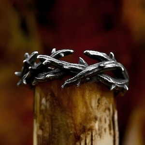 May include: A silver-colored ring with a thorny design. The ring is made of metal and has a detailed, textured appearance. The thorns are sharp and point outwards, creating a crown-like shape. The ring is displayed on a wooden surface.