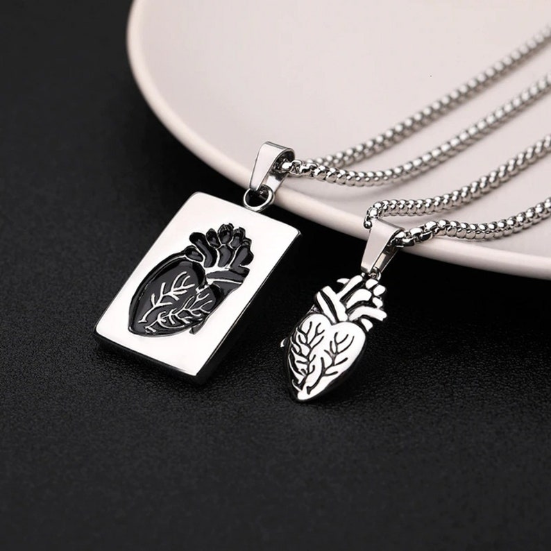 Pu&ograve; includere: Due collane d'argento con ciondoli a forma di cuore anatomico. Un ciondolo &egrave; di forma rettangolare con un cuore nero all'interno. L'altro ciondolo &egrave; a forma di cuore con un cuore nero all'interno.