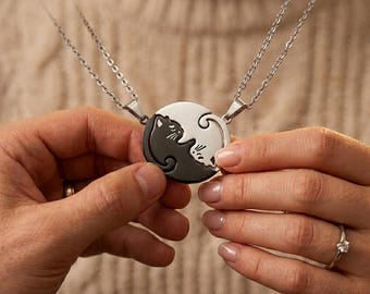 Può includere: Due collane d'argento con ciondoli abbinati. Ogni ciondolo presenta un design Yin e Yang con una silhouette di gatto in bianco e nero. Le collane sono tenute in mano, evidenziando i dettagli dei ciondoli e la catena d'argento.