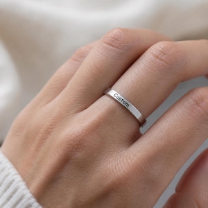 Könnte beinhalten: Ein silberner Ring mit dem Wort "Custom" auf dem Band eingraviert. Der Ring wird an einem Finger gezeigt, mit einem weichen Hintergrund.