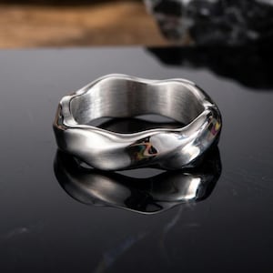 May include: A silver-colored ring with a wavy, organic design. The ring has a smooth, polished exterior and a brushed interior. The ring is displayed on a reflective black surface.