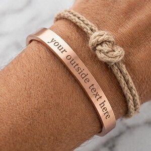 Può includere: Un bracciale a polsino color rame con la scritta incisa "your outside text here" è mostrato. È visibile anche un secondo braccialetto in corda beige annodata. I braccialetti sono indossati al polso di una persona.