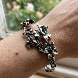 May include: Silver-toned floral cuff bracelet. The bracelet features detailed flower and leaf designs. The cuff is worn on a wrist, with a blurred background of a window and foliage.