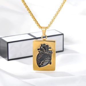 Pu&ograve; includere: Una collana con catena dorata con un ciondolo rettangolare che presenta un design di cuore anatomico nero.