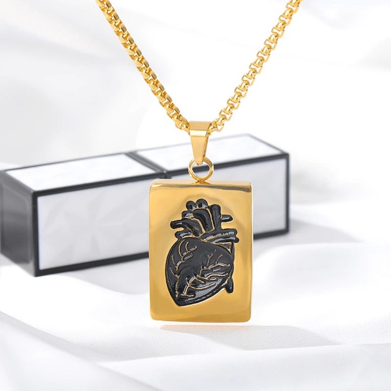 Pu&ograve; includere: Una collana con catena dorata con un ciondolo rettangolare che presenta un design di cuore anatomico nero.