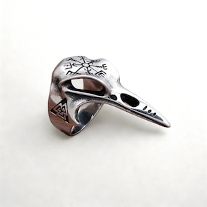 Op de afbeelding: Een zilverkleurige ring in de vorm van een vogel schedel. De ring heeft een gedetailleerd ontwerp van een raven schedel met een puntige snavel en oogkassen. De bovenkant van de ring heeft een kompasachtig symbool en de zijkant heeft een driehoekig symbool.