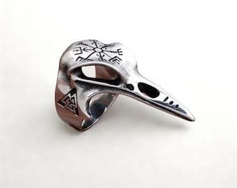Puede incluir: Un anillo plateado con forma de cráneo de pájaro. El anillo presenta un diseño detallado de un cráneo de cuervo con pico puntiagudo y cuencas oculares. La parte superior del anillo tiene un símbolo similar a una brújula y el lateral un símbolo triangular.