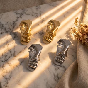 May include: Two pairs of earrings, one gold-toned and one silver-toned, each featuring a sculpted, abstract human face design with prominent facial features and horizontal lines across the lower portion, presented as four individual pieces.