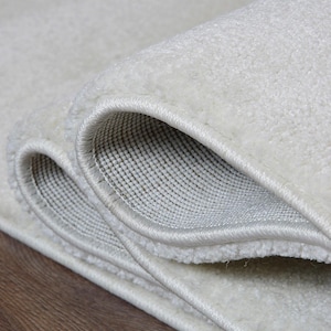 May include: Close-up of a rolled-up, off-white rug. The rug has a soft, textured surface and a light-colored border. The underside of the rug reveals a woven backing. The rug is placed on a wooden surface.