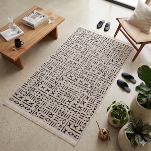 Modern boho mudcloth area rug with black tribal geometric pattern on ivory background and fringe, styled in a minimalist Scandinavian living room.