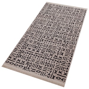 May include: A beige and black patterned rug with a fringe border. The pattern is geometric and abstract, featuring lines, dots, and crosses.
