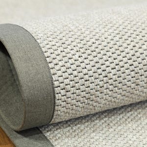 May include: Close-up of a rolled-up, neutral-toned rug with a woven texture. The rug features a gray border that frames the woven pattern. The rug's design is simple and modern, suitable for various interior styles.
